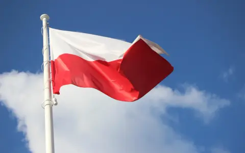 Biało-czerwona bandera polska powiewa na maszcie na tle błękitnego nieba z chmurami.
