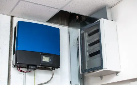 Niebieski falownik, kluczowy element instalacji fotowoltaicznej, zamontowany obok białej skrzynki z zabezpieczeniami.