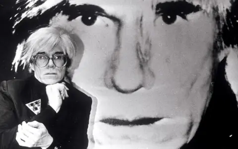 Andy Warhol, Ikone der Pop Art, posiert vor einem riesigen Porträt von sich selbst. Seine Kunstrichtung prägte die moderne Kunst.