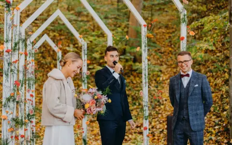 Piękna panna młoda z bukietem, pan młody przemawia do mikrofonu, a pan młody z uśmiechem. Wesele jesienią w otoczeniu kolorowych liści.
