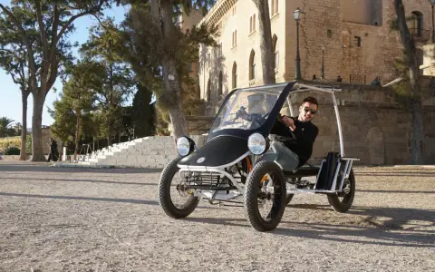 Ein Mann fährt einen Duo Moke durch eine sonnige Gasse mit historischen Gebäuden im Hintergrund.