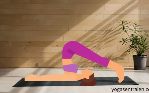 En person utför plogen yoga på en matta framför en trävägg.