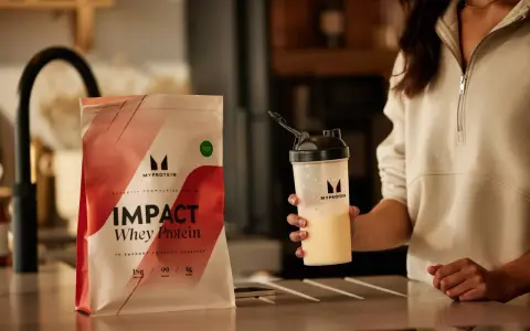 Eine Person hält einen Shaker mit einem cremigen Getränk. Daneben liegt eine Packung Myprotein Impact Whey Protein. Perfekt für den proteinshake vor dem schlafen.