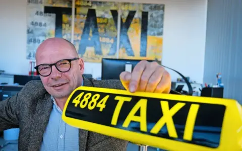 Mann mit Brille hält ein gelbes Taxi-Schild mit der Nummer 4884. Im Hintergrund Kunstwerke mit "TAXI" und der Zahl 1884.