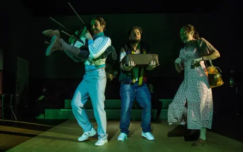 Iwo Orłowski życiorys: scena teatralna z mężczyzną niosącym krzesło i walizkę, obok kobieta w sukience.