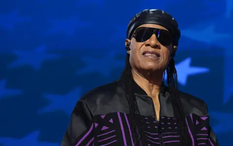 Stevie Wonder heute, mit Sonnenbrille und schwarzer Kappe, trägt eine Jacke mit lila Muster vor blauem Sternenhintergrund.