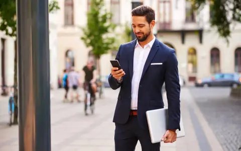 Mężczyzna w eleganckim stroju biznesowym męskim sprawdza telefon, trzymając laptopa.