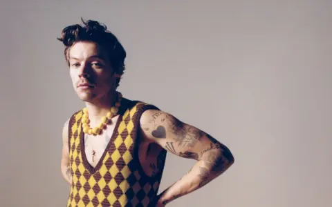 Harry Styles w kamizelce w romby i żółtym naszyjniku, jakby inspirował nowe harry styles utwory.