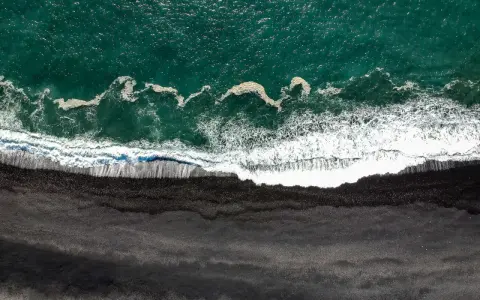 Gran Canaria: Türkisfarbenes Meer trifft auf einen dramatischen schwarzen Strand. Wellen brechen sich auf dem dunklen Sand.