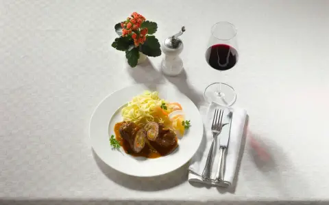 Ein Teller Rouladen mit Bandnudeln und einem Glas feinherb Wein. Dazu ein kleines Blümchen und Besteck.