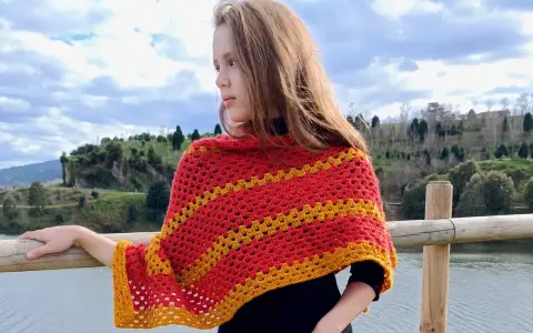 Chica con un pañuelo triangular crochet rojo y amarillo, apoyada en una barandilla de madera junto a un lago.