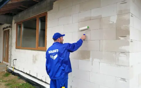 Pracownik w niebieskim kombinezonie nakłada wałkiem grunt na ścianę z bloczków. To kluczowy etap przygotowania pod styropian.