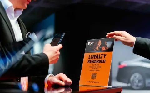 Ein Mann hält ein Smartphone, während eine Hand ein orangefarbenes Schild mit der Aufschrift "Loyalty Rewarded" und dem SIXT ONE Logo hochhält.