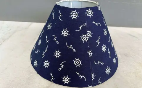 Un abat-jour conique bleu marine orné de motifs d'ancre et de roue de gouvernail. Idéal pour fabriquer un abat-jour en tissu plissé avec une touche marine.
