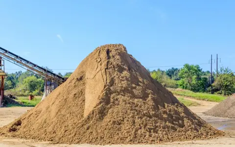 Ogromna hałda piasku na placu budowy, zastanawiasz się, ile kosztuje tona piasku? W tle taśmociąg i drzewa.