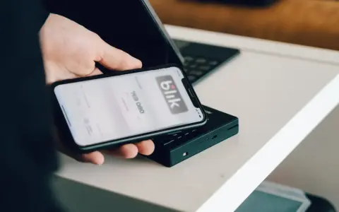 Dłoń trzyma smartfon z aplikacją Blik i kodem transakcji nad terminalem płatniczym.