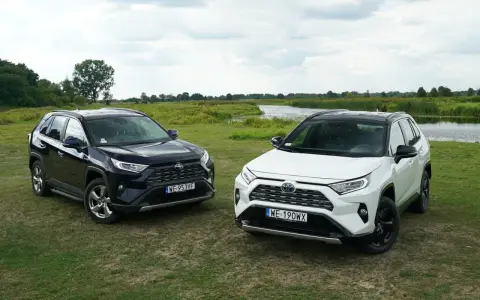 Dwa SUV-y: ciemny i biały Toyota RAV4 hybryda, stoją na łące nad jeziorem.