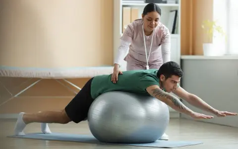 Fizjoterapeutka wspiera mężczyznę w ćwiczeniach na piłce. Rekonwalescencja po nietrzymaniu moczu, przygotowanie do powrotu do pracy fizycznej.