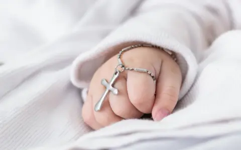 Dziecięca dłoń trzyma krzyżyk na łańcuszku, symbol tego, co jest potrzebne do chrztu.