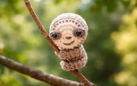 Adorable perezoso amigurumi de crochet, con grandes ojos y una sonrisa, aferrado a una rama.