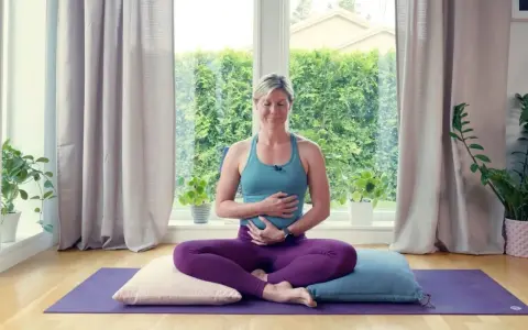 Kvinna i lila kläder sitter i meditationsställning på en yogamatta, med händerna över magen. Hon utövar yoga gravid.