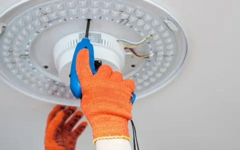 Ręka w pomarańczowej rękawicy odkręca śrubę plafonu LED. Instrukcja jak zdjąć plafon krok po kroku.