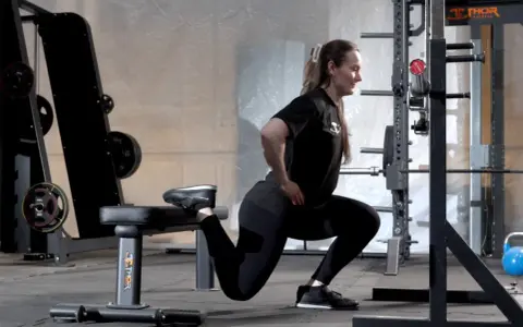 Kvinna utför en split squat i ett gym. Hennes ena fot vilar på en bänk bakom henne.