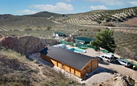 Casa de madera con piscina y caravana en terreno rústico, rodeada de olivos y colinas. Ideal para quienes buscan un refugio natural.