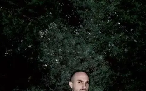 Un hombre calvo, con mirada intensa, posa frente a un fondo de follaje oscuro. Podría ser un sicario rapero, listo para soltar rimas letales.