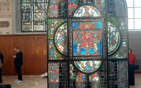 Un vitrail coloré, représentant des scènes bibliques, se dresse dans un intérieur. Un homme consulte son téléphone devant ce chef-d'œuvre.