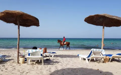 Piękna plaża na Djerbie, Tunezja. Jeździec na koniu przemierza brzeg morza, podczas gdy inni odpoczywają pod parasolami.
