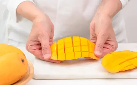 Ręce trzymają pokrojone w kostkę mango, pokazując, jak pokroić mango. Obok leżą całe owoce.