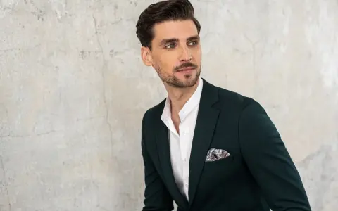 Elegancki mężczyzna w ciemnozielonej marynarce i białej koszuli z ozdobną poszetką. Jego kołnierzyk koszuli, czyli stójka, jest lekko rozpięty.