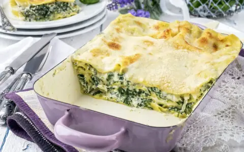 Pyszne lasagne ze szpinakiem w fioletowym naczyniu, gotowe do podania. Obok talerze z porcją i sztućce.