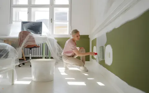 Kobieta maluje ścianę na zielono, klęcząc obok wiadra z farbą.