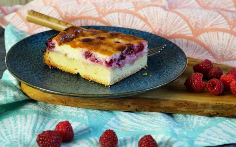 Ein Stück Himbeer Schmand Kuchen auf einem blauen Teller, daneben frische Himbeeren.