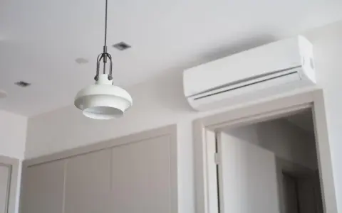 Biała klimatyzacja ścienna nad drzwiami, obok lampy wiszącej.
