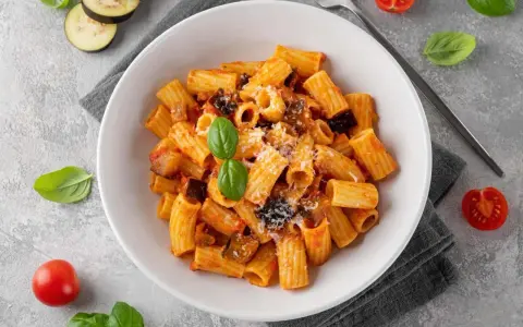 En skål med pasta alla norma, toppad med basilika och riven ost. Tomater och aubergineskivor ligger bredvid.