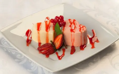 Dwie panna cotta w kształcie serc, polane sosem truskawkowym, ozdobione owocami i miętą.