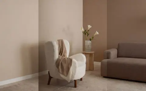 Gemütliches Wohnzimmer mit beigen und braunen Wandfarben. Ein flauschiger Sessel mit Decke und ein Sofa laden zum Entspannen ein.