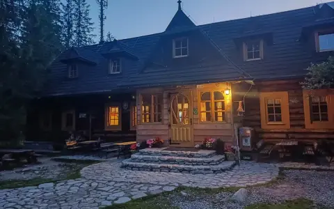 Drewniana chata w górach, oświetlona ciepłym światłem, to idealna baza pod roztokami. Kamienna ścieżka prowadzi do wejścia.