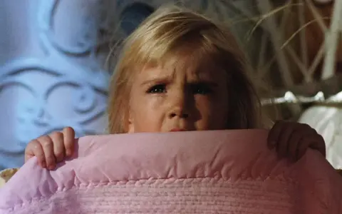 Bambina spaventata si nasconde sotto una coperta rosa, gli occhi sgranati in un'espressione di puro horror psicologico.