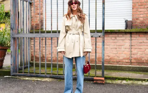 Mujer con gabardina beige y vaqueros bootcut, gafas rojas y bolso rojo.