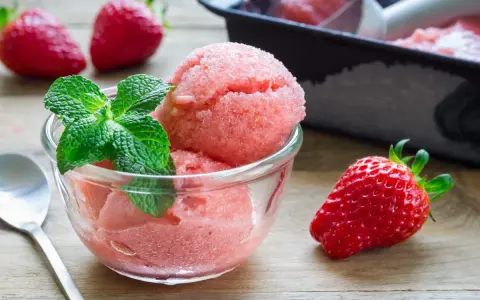 Erfrischendes Erdbeer-Sorbet-Eis in einer Glasschale, garniert mit Minze. Daneben liegen frische Erdbeeren.