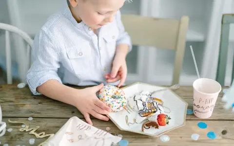 En pojke med en randig skjorta tar en donut med strössel. Det är en festlig dukning med konfetti och en mugg med texten "Hip Hip Hooray". En härlig stund av burken lek.