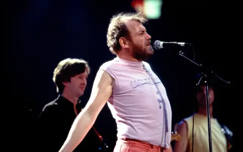 Joe Cocker śpiewa na scenie, a jego charakterystyczne utwory niosą się po sali. W tle widać innych muzyków.