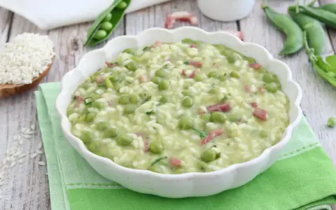Un cremoso piatto di risi e bisi, con piselli freschi e cubetti di pancetta, servito in una ciotola bianca su un tovagliolo verde.