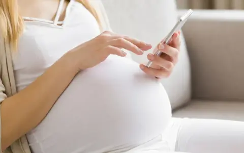 Kobieta w ciąży, w białej bluzce i kardiganie, siedzi na kanapie i korzysta z telefonu. Może sprawdzać zwolnienie lekarskie w ciąży.