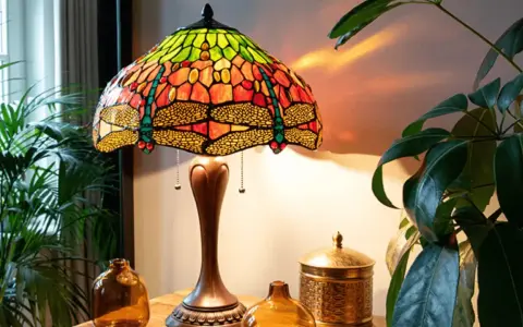 Lampe tiffany origine, avec abat-jour en vitrail coloré orné de libellules, posée sur une table en bois près de plantes vertes.