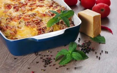 Pyszna zapiekanka makaronowa z kiełbasą, zapieczona pod złotym serem. Obok kawałek sera, pomidory i przyprawy.